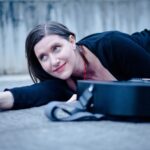 Eine Frau dunkel gekleidet mit roter Kette liegt mit ihrer Gitarre am Boden