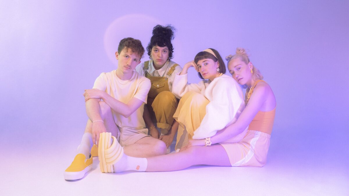 Ein Mann und drei Frauen sitzen in orangener Kleidung vor violettem Hintergrund