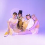 Ein Mann und drei Frauen sitzen in orangener Kleidung vor violettem Hintergrund