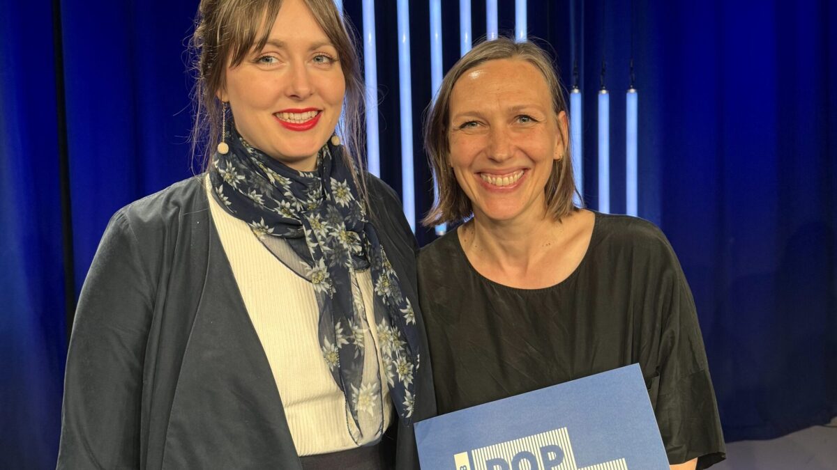 Zwei Frauen stehen auf einer blau beleuchteten Bühne und lachen in die Kamera. Eine Frau hält eine Urkunde mit der Aufschrift: Wettbewerb Pop Musik Sachsen 2024.