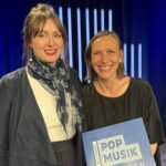 Zwei Frauen stehen auf einer blau beleuchteten Bühne und lachen in die Kamera. Eine Frau hält eine Urkunde mit der Aufschrift: Wettbewerb Pop Musik Sachsen 2024.