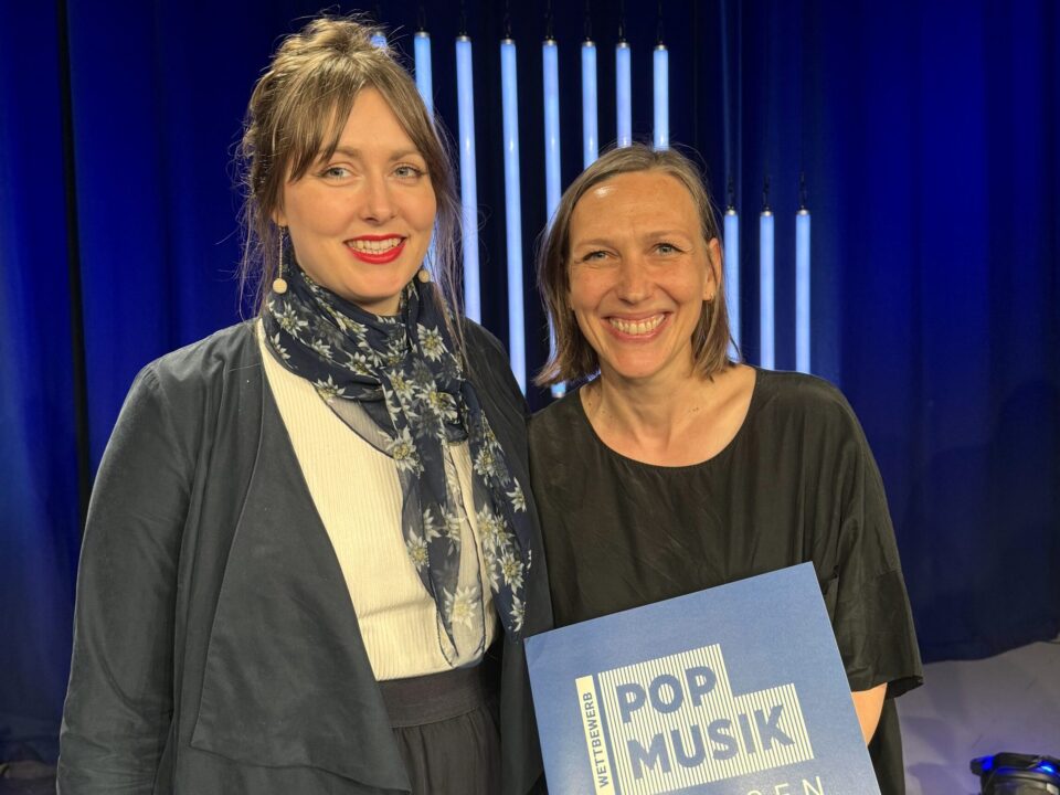 Zwei Frauen stehen auf einer blau beleuchteten Bühne und lachen in die Kamera. Eine Frau hält eine Urkunde mit der Aufschrift: Wettbewerb Pop Musik Sachsen 2024.