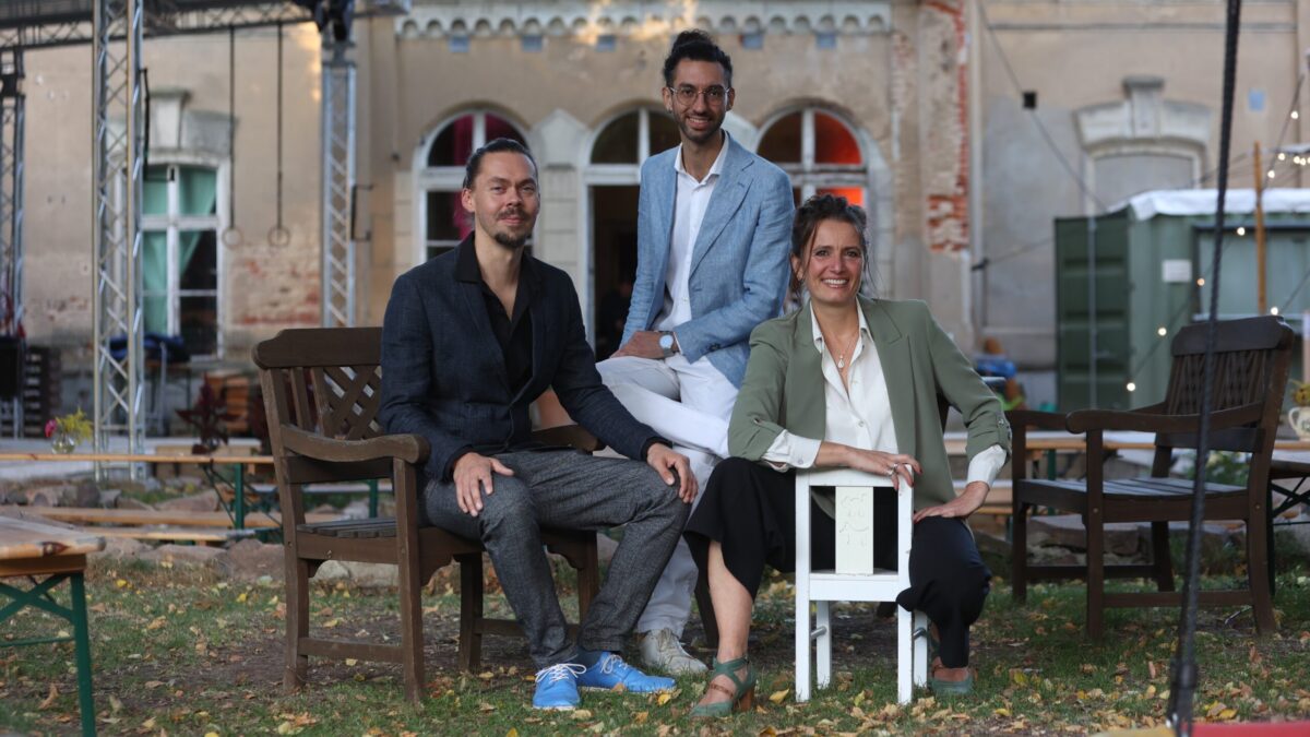 Zwei Männer und eine Frau sitzen auf Stühlen vor einem alten Gebäude, alle lächeln
