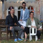 Zwei Männer und eine Frau sitzen auf Stühlen vor einem alten Gebäude, alle lächeln