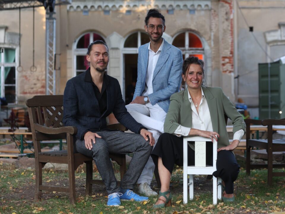 Zwei Männer und eine Frau sitzen auf Stühlen vor einem alten Gebäude, alle lächeln