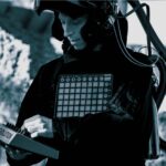 Ein schwarz-weißes Bild. Zu sehen ist ein weißer Mann mit einem Synthesizer in der Hand und einem Helm mit Schlauch auf dem Kopf