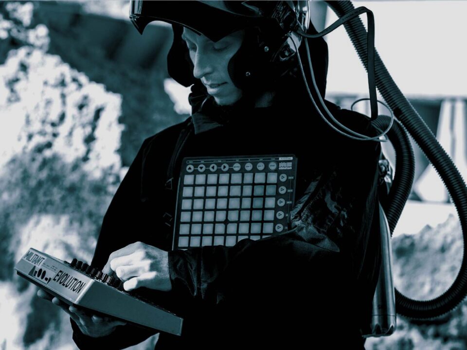 Ein schwarz-weißes Bild. Zu sehen ist ein weißer Mann mit einem Synthesizer in der Hand und einem Helm mit Schlauch auf dem Kopf