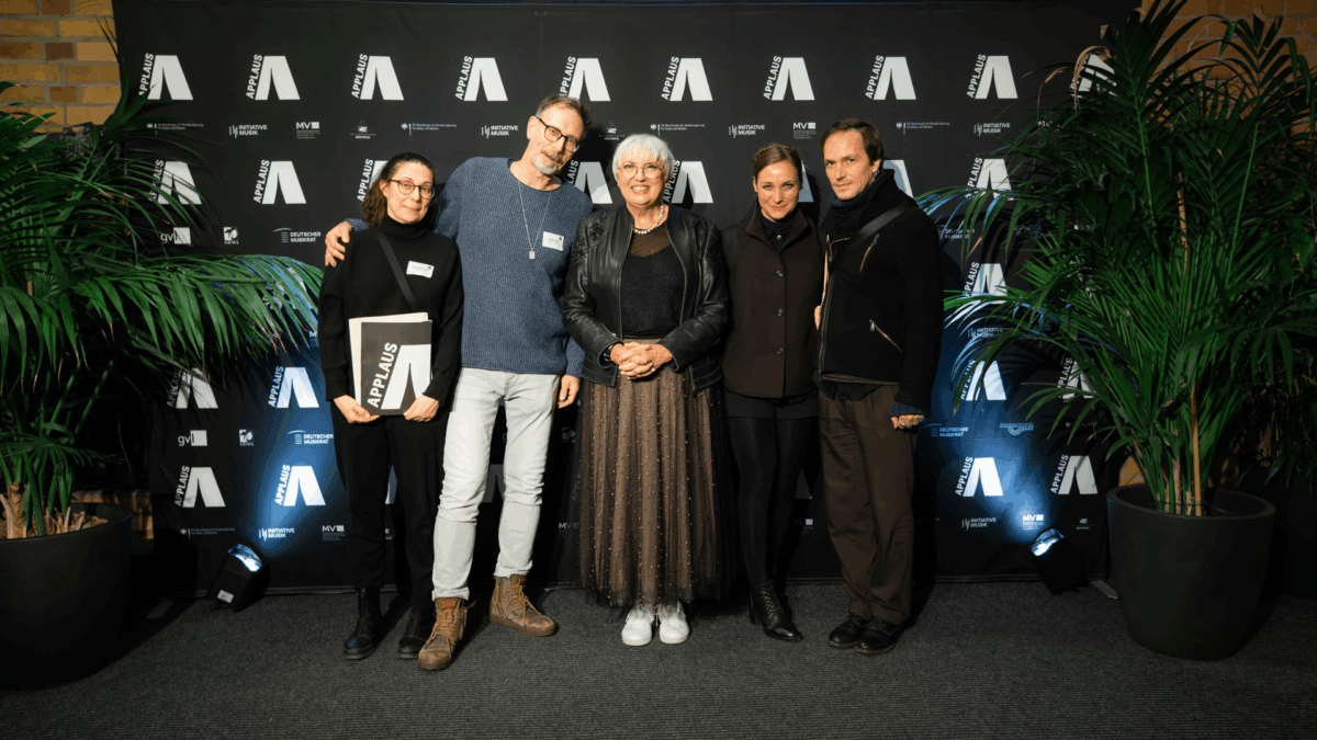 Eine Gruppe von fünf weißen Menschen, zwei Männer und eine Frau. Die Frau links hält eine Urkunde in der Hand mit der Aufschrift APPLAUS. In der Mitte steht Kulturministerin Claudia Roth.