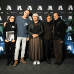 Eine Gruppe von fünf weißen Menschen, zwei Männer und eine Frau. Die Frau links hält eine Urkunde in der Hand mit der Aufschrift APPLAUS. In der Mitte steht Kulturministerin Claudia Roth.