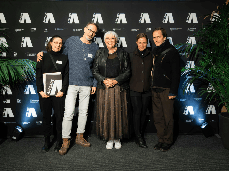 Eine Gruppe von fünf weißen Menschen, zwei Männer und eine Frau. Die Frau links hält eine Urkunde in der Hand mit der Aufschrift APPLAUS. In der Mitte steht Kulturministerin Claudia Roth.
