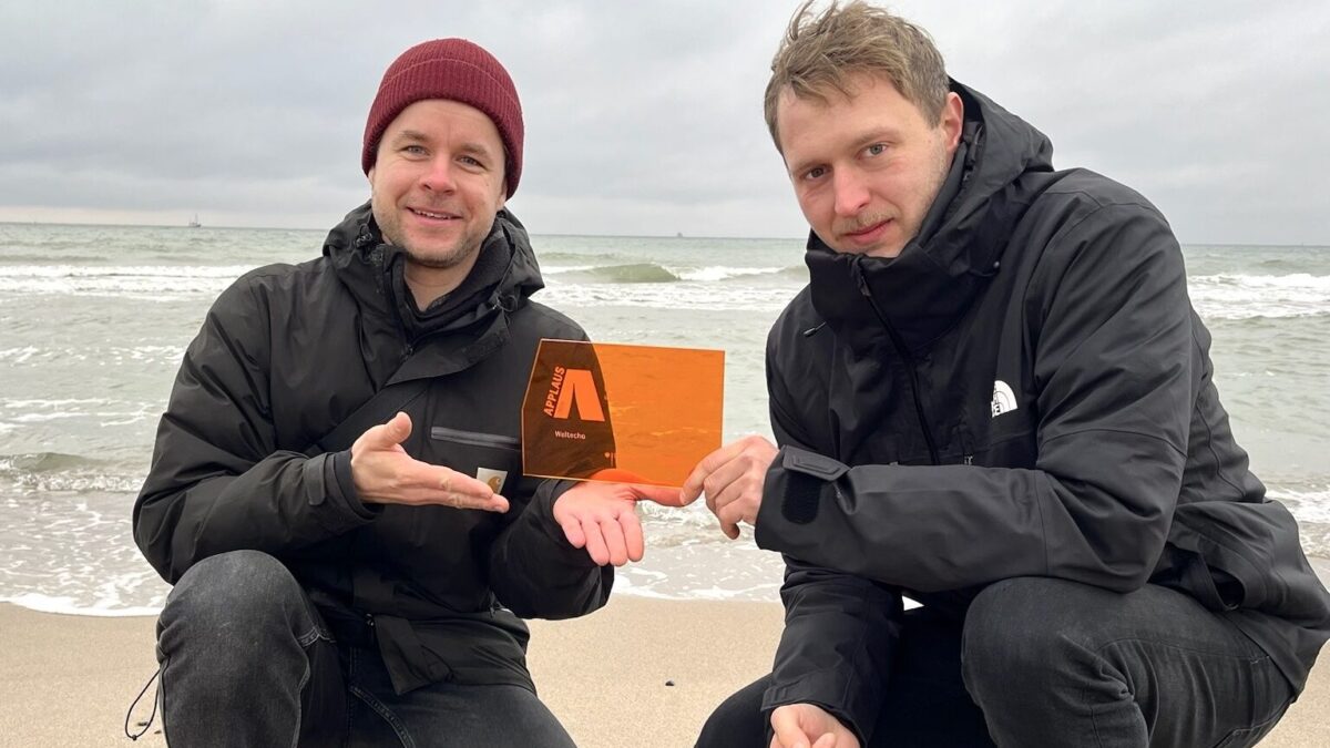 Zwei weiße Männer in der Hocke an einem Strand, sie halten gemeinsam einen orangenen Preis aus Glas in der Hand mit der Aufschrift APPLAUS.