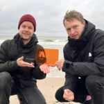 Zwei weiße Männer in der Hocke an einem Strand, sie halten gemeinsam einen orangenen Preis aus Glas in der Hand mit der Aufschrift APPLAUS.