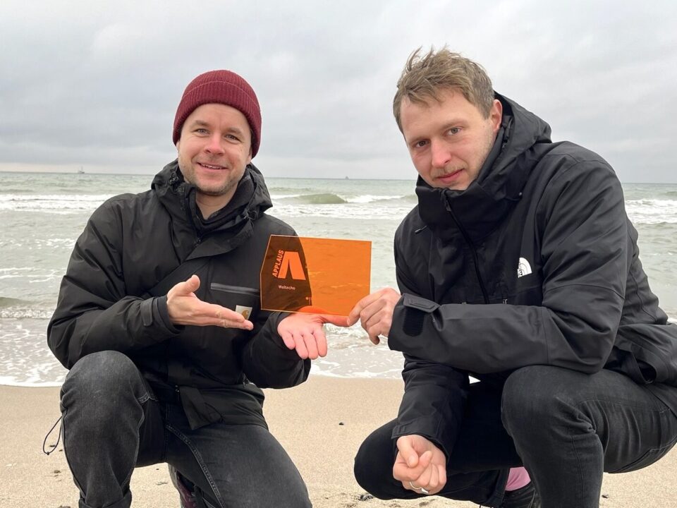 Zwei weiße Männer in der Hocke an einem Strand, sie halten gemeinsam einen orangenen Preis aus Glas in der Hand mit der Aufschrift APPLAUS.