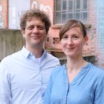 Zwei weiße Personen vor einer groben Mauer. Links ein Mann mit Locken und weißem Hemd, rechts eine Frau mit Seitenscheitel und Jeansbluse. Beide lächeln.