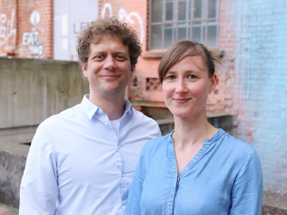 Zwei weiße Personen vor einer groben Mauer. Links ein Mann mit Locken und weißem Hemd, rechts eine Frau mit Seitenscheitel und Jeansbluse. Beide lächeln.