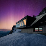 Eine Hütte mit großer Fensterfront im Schnee, im Hintergrund lila weiße Nordlichter