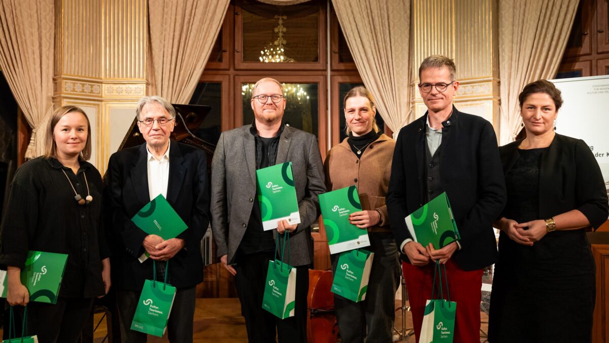 Sechs weiße Menschen, drei Frauen und drei Männer, alle schick gekleidet. Fünf Menschen halten eine grüne Urkunde und grüne Taschen in der Hand mit der Aufschrift: Kultur Tourismus Sachsen.