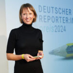 Eine Frau mit schulterlangen dunkelblonden Haaren in schwarz gekleidet vor einer Wand mit der Aufschrift: Deutscher Reporter:innenpreis 2024