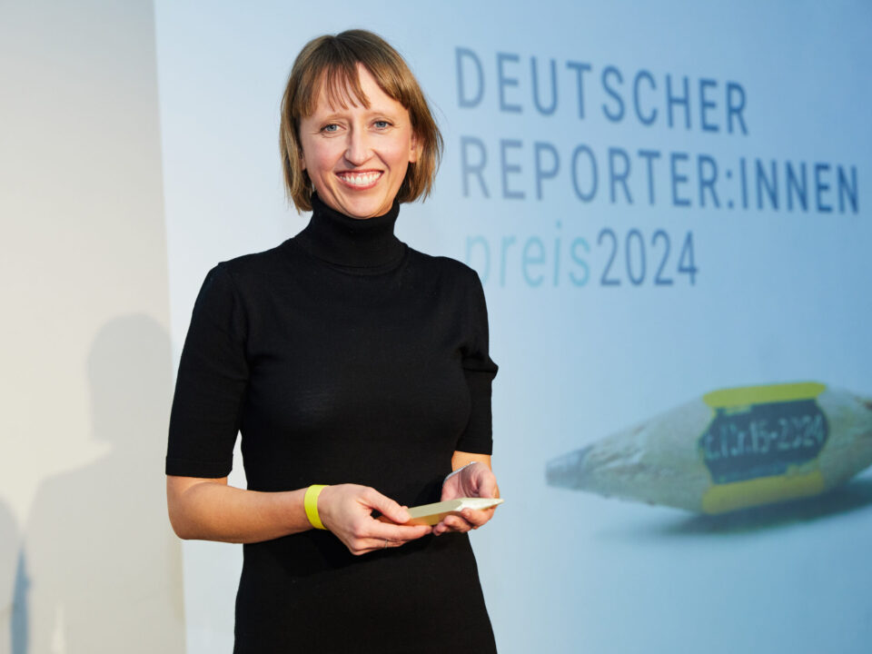 Eine Frau mit schulterlangen dunkelblonden Haaren in schwarz gekleidet vor einer Wand mit der Aufschrift: Deutscher Reporter:innenpreis 2024