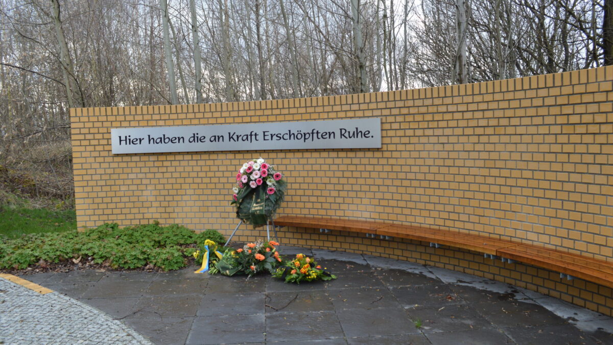 Eine Mauer mit langer Bank, darauf ein Kranz und die Aufschrift: Hier haben die an Kraft erschöpften Ruhe.