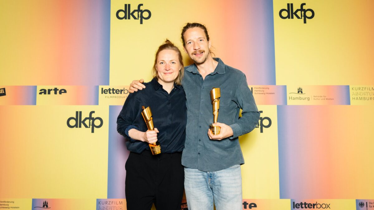 Zwei weiße Personen vor einer orang-gelben Wand, Frau und Mann. Aufschrift: dkfp. Sie halten beide einen Pokal in der Hand und lächeln.
