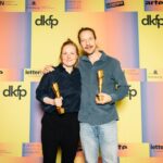 Zwei weiße Personen vor einer orang-gelben Wand, Frau und Mann. Aufschrift: dkfp. Sie halten beide einen Pokal in der Hand und lächeln.