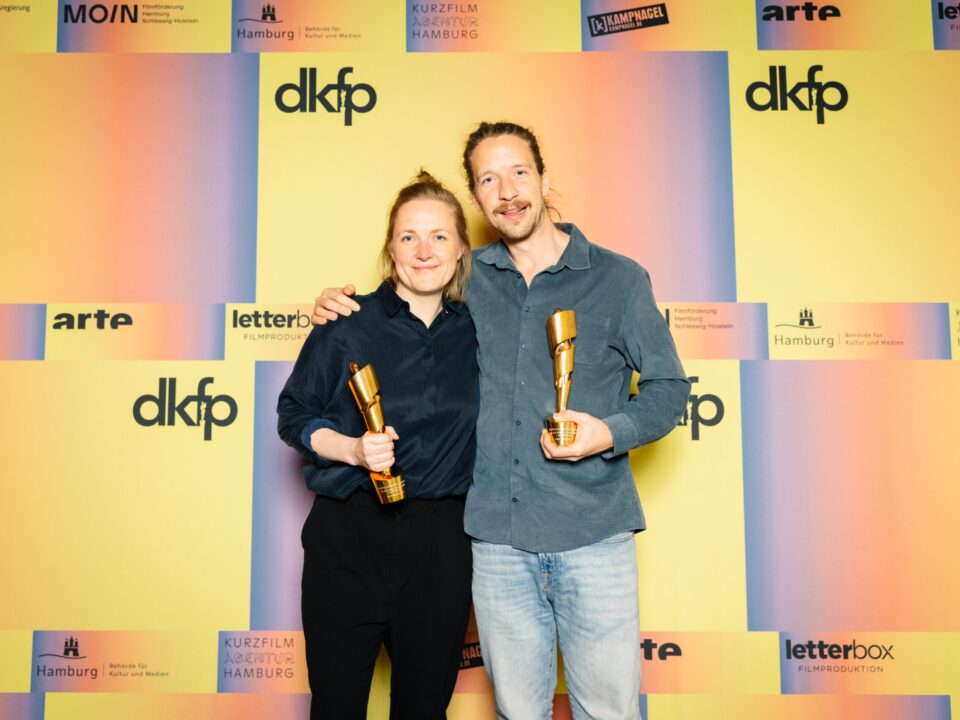 Zwei weiße Personen vor einer orang-gelben Wand, Frau und Mann. Aufschrift: dkfp. Sie halten beide einen Pokal in der Hand und lächeln.