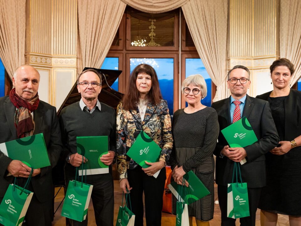 Sechs weiße Personen, drei Frauen, drei Männer, alle schick gekleidet. Fünf Menschen halten eine grüne Urkunde und grüne Taschen in der Hand mit der Aufschrift: Kultur Tourismus Sachsen.
