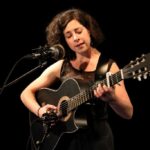 Eine weiße Frau in schwarzer Kleidung vor schwarzem Hintergrund, sie trägt eine Gitarre in den Händen und singt.