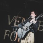 Eine weiße Frau in schwarzer Kleidung mit beiger Hose vor schwarzem Hintergrund, sie trägt eine Gitarre in den Händen und singt.