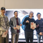Vier weiße junge Männer mit Mütze vor einer leuchtenden Wand, in der Hand halten drei von ihnen eine Urkunde mit der Aufschrift EMIL.