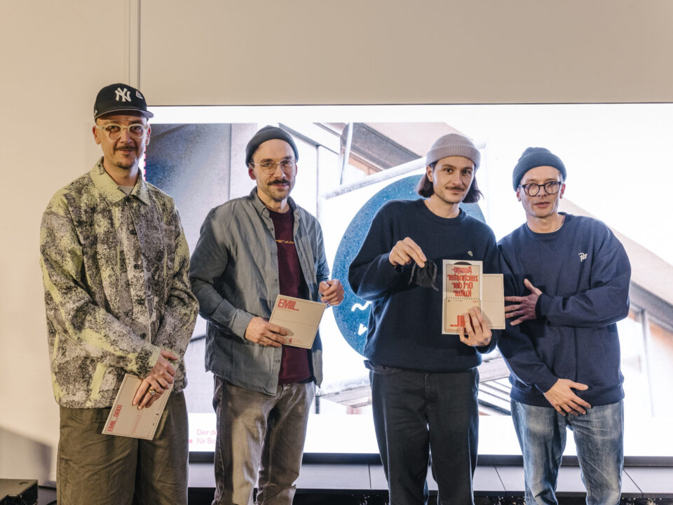 Vier weiße junge Männer mit Mütze vor einer leuchtenden Wand, in der Hand halten drei von ihnen eine Urkunde mit der Aufschrift EMIL.