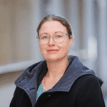 Weiße Frau mit Brille und zusammengebundenen haaren, sie trägt eine dunkleJacke