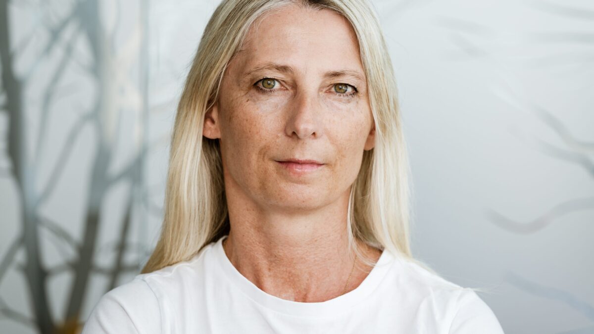 Blonde Frau mittleren Alters in einem weißen T-Shirt, im Hintergrund Nebel und kahle Bäume.