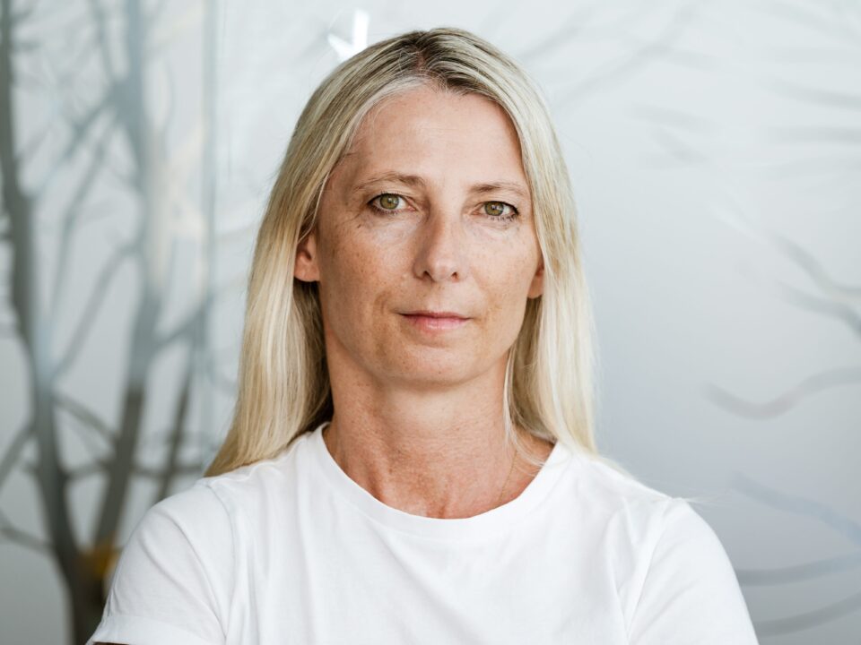 Blonde Frau mittleren Alters in einem weißen T-Shirt, im Hintergrund Nebel und kahle Bäume.
