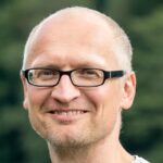 Lächelnder weißer Mann mit gräulicher Halbglatze und schwarzer Brille, im Hintergrund verschwommene Hecke