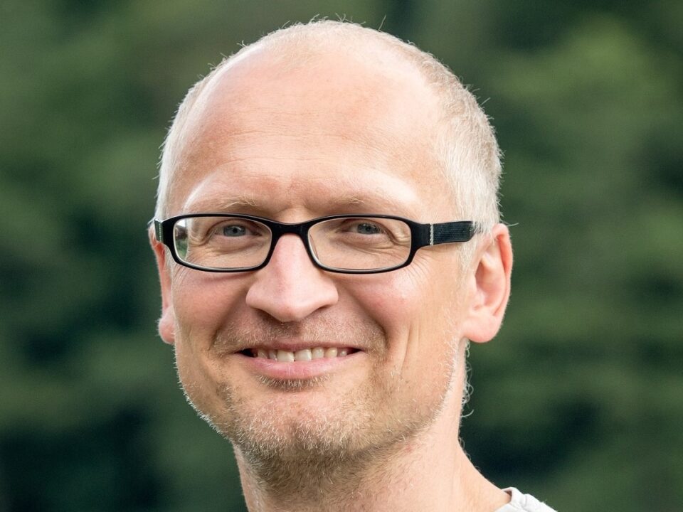 Lächelnder weißer Mann mit gräulicher Halbglatze und schwarzer Brille, im Hintergrund verschwommene Hecke
