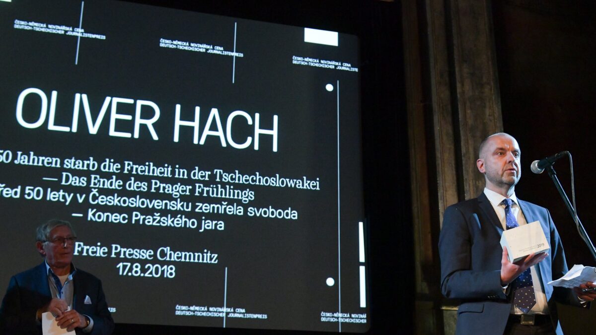 Ein weißer Mann mittleren Alters mit abrasierten Haaren und Teitagebart im Anzug, er hält einen Preis in der Hand mit der Aufschrift: 2019. Im Hintergrund ein großer Schriftzug: Oliver Hach, das Ende des Prager Frühlings.