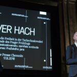 Ein weißer Mann mittleren Alters mit abrasierten Haaren und Teitagebart im Anzug, er hält einen Preis in der Hand mit der Aufschrift: 2019. Im Hintergrund ein großer Schriftzug: Oliver Hach, das Ende des Prager Frühlings.